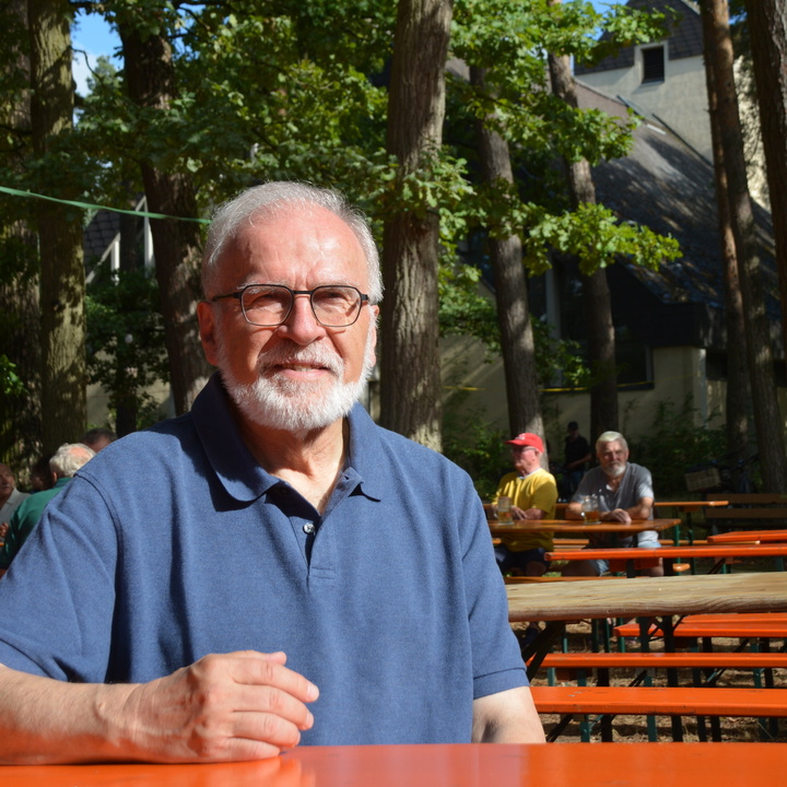 Pfarrer Gregor Mühleck sitzt an einer Biertisch-Garnitur auf dem Vorplatz der Kirche St. Hedwig. Im Hintergrund sind hohe Bäume zu sehen, die die Kirche fast verdecken. Zudem machen es sich erste Gäste beim Pfarrfest bequem.