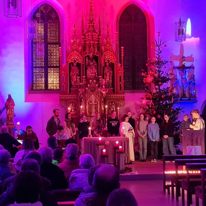 Blick auf den Altarraum der Kirche. Die Kirche ist mit Strahlern lila und rosa erleuchtet, im Altarraum stehen einige Jugendliche und lesen etwas vor.