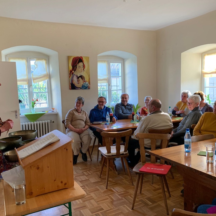 Links im Bild steht eine Frau an einem Tisch, auf dem mehrere Klangschalen stehen, und erzählt etwas. Rechts im Bild sieht man mehrere Leute, die an Tischen sitzen und aufmerksam zuhören.