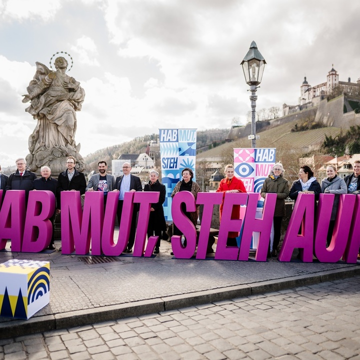 Auf der Mainbrücke stehen Menschen hinter dem Schriftzug "Hab Mut Steh Auf". Im Hintergrund ist die Festung Marienberg zu sehen.