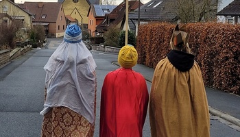 Blick von hinten auf drei Sternsinger, der mittlere hält einen Stern hoch. Im Hintergrund sieht man die Kirche.